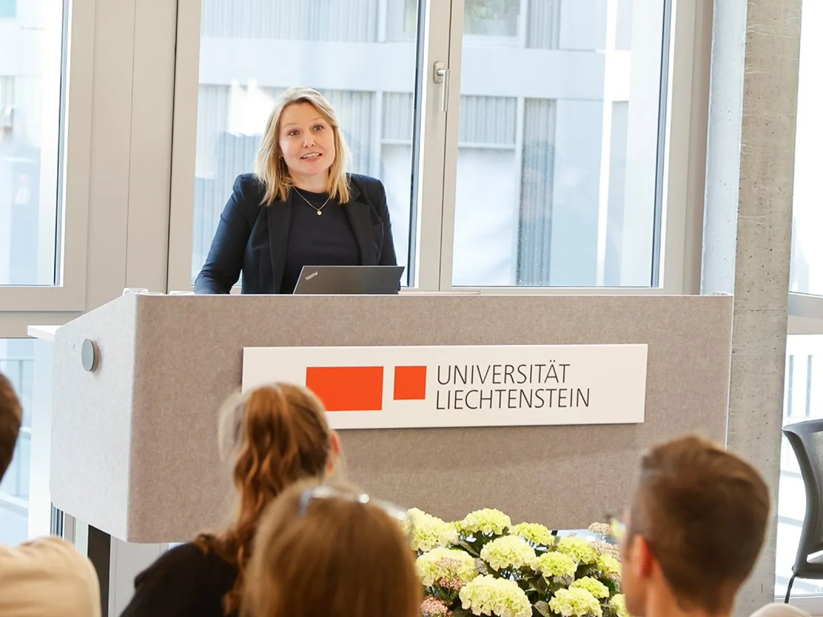 Eine Person steht an einem Rednerpult der Universität Liechtenstein und spricht zu einem Publikum. Im Hintergrund sind große Fenster zu sehen. Vor dem Pult sind Blumen arrangiert.