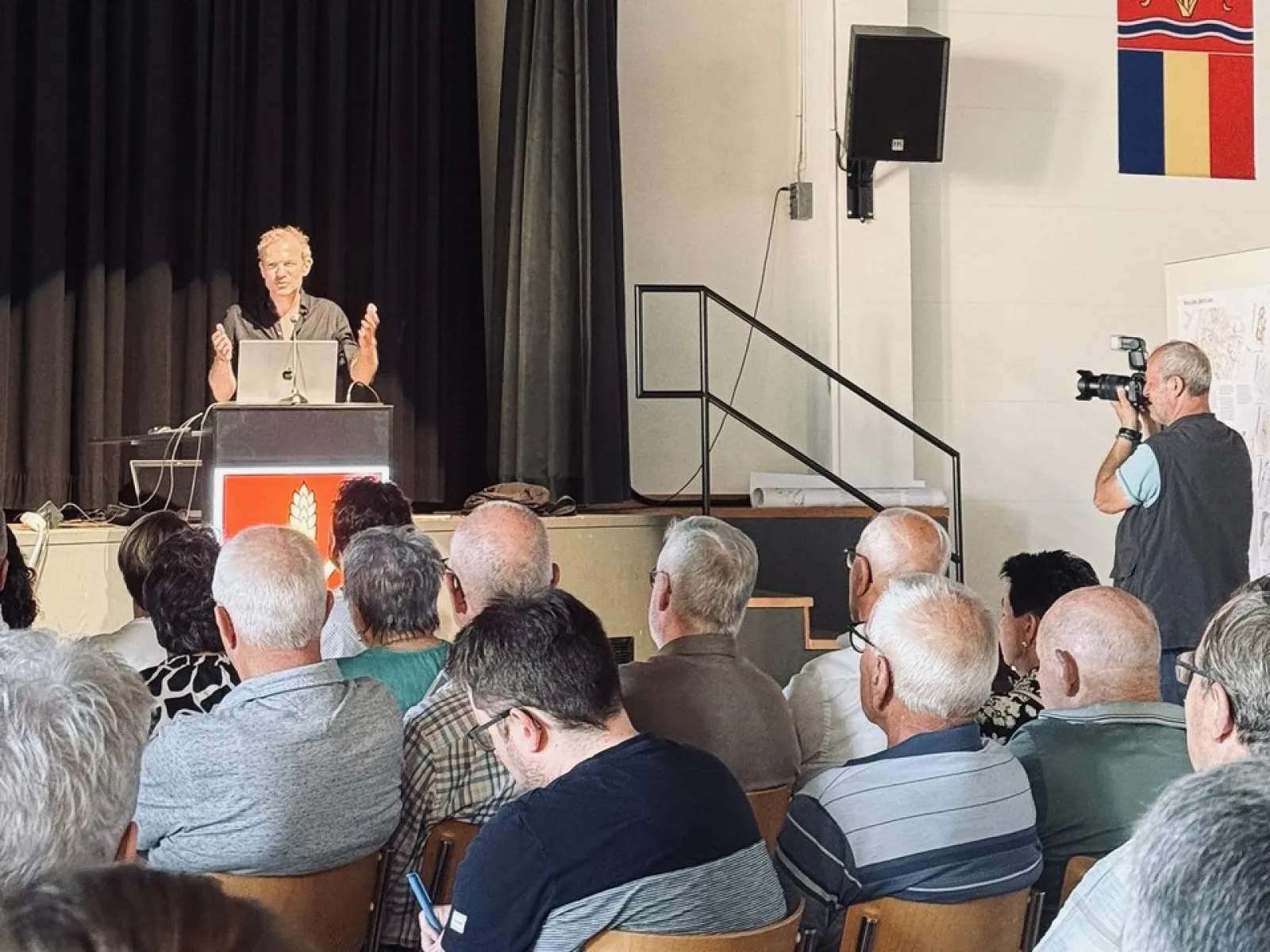 Ein Vortragender steht an einem Rednerpult vor einem Publikum in einem Saal. Im Hintergrund sind schwarze Vorhänge und ein Fotograf macht Bilder. An der Wand hängt ein Wappen.