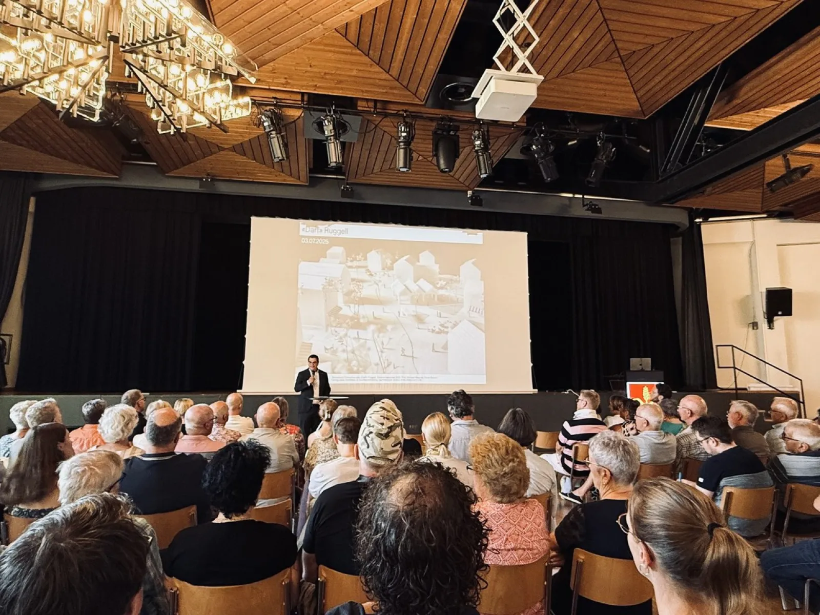 Eine Person hält einen Vortrag vor einem Publikum in einem Saal. Auf der Bühne ist eine große Leinwand mit einer Präsentation zu sehen. Der Raum hat eine moderne Holzdecke mit Beleuchtung.