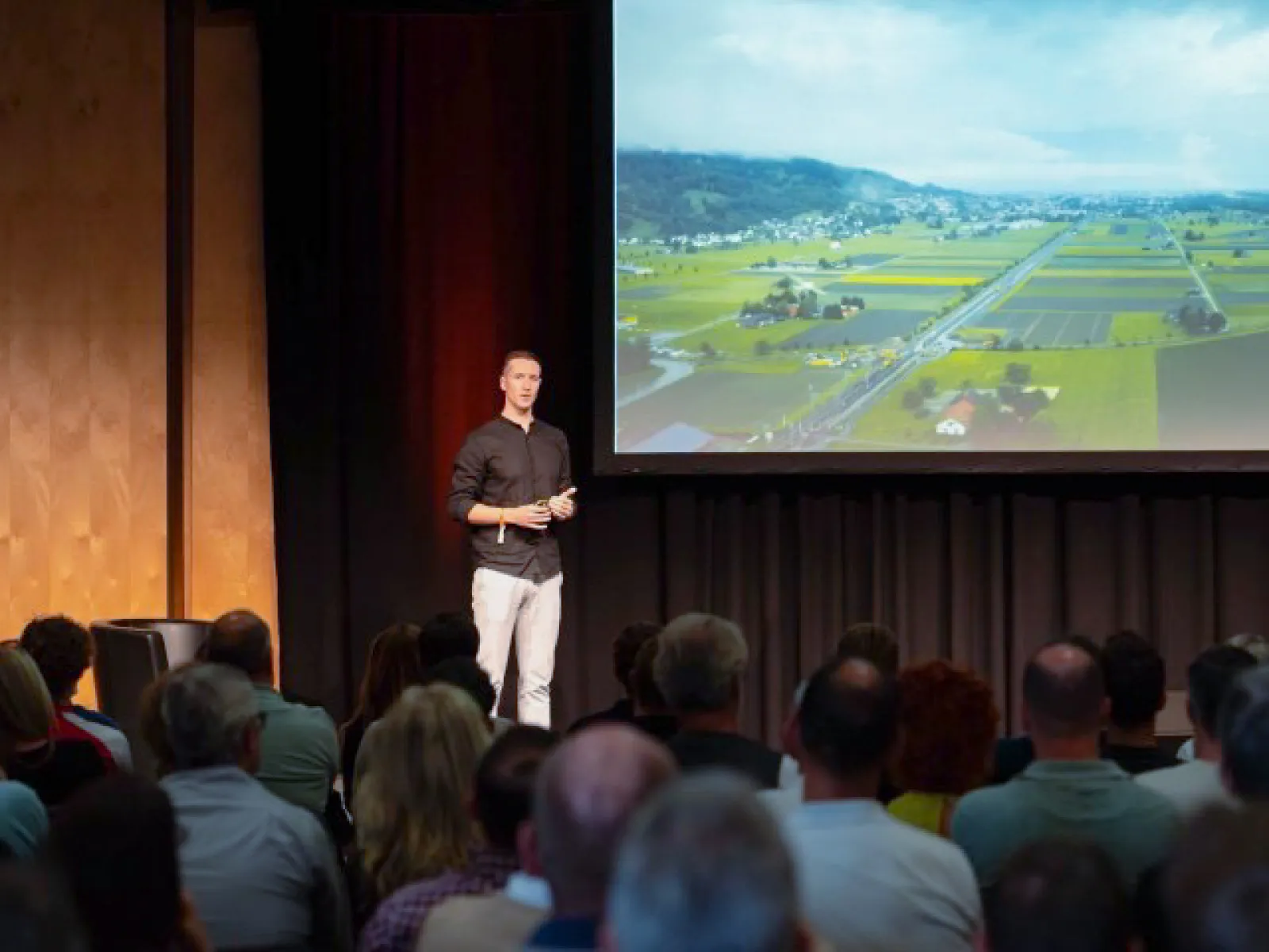 Speaker präsentiert auf Bühne vor grosser Leinwand mit Landschaftsbild während Fachvortrag