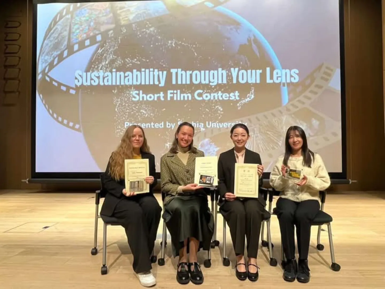 Vier weibliche Personen sitzen auf einem Stuhl und halten ein Zertifikat in der Hand