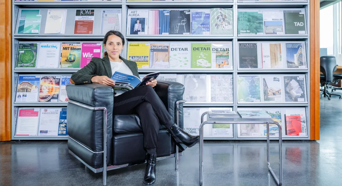 Frau sitzt entspannt auf einem schwarzen Ledersessel in der Bibliothek der Universität Liechtenstein und liest in einer Fachzeitschrift, im Hintergrund ein Regal mit bunten Magazincovern.