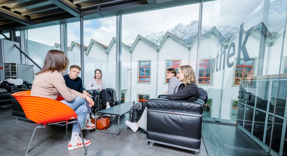 Studierende der Universität Liechtenstein sitzen in einer gemütlichen Sitzecke mit Blick auf die Alpen und tauschen sich in entspannter Atmosphäre aus.