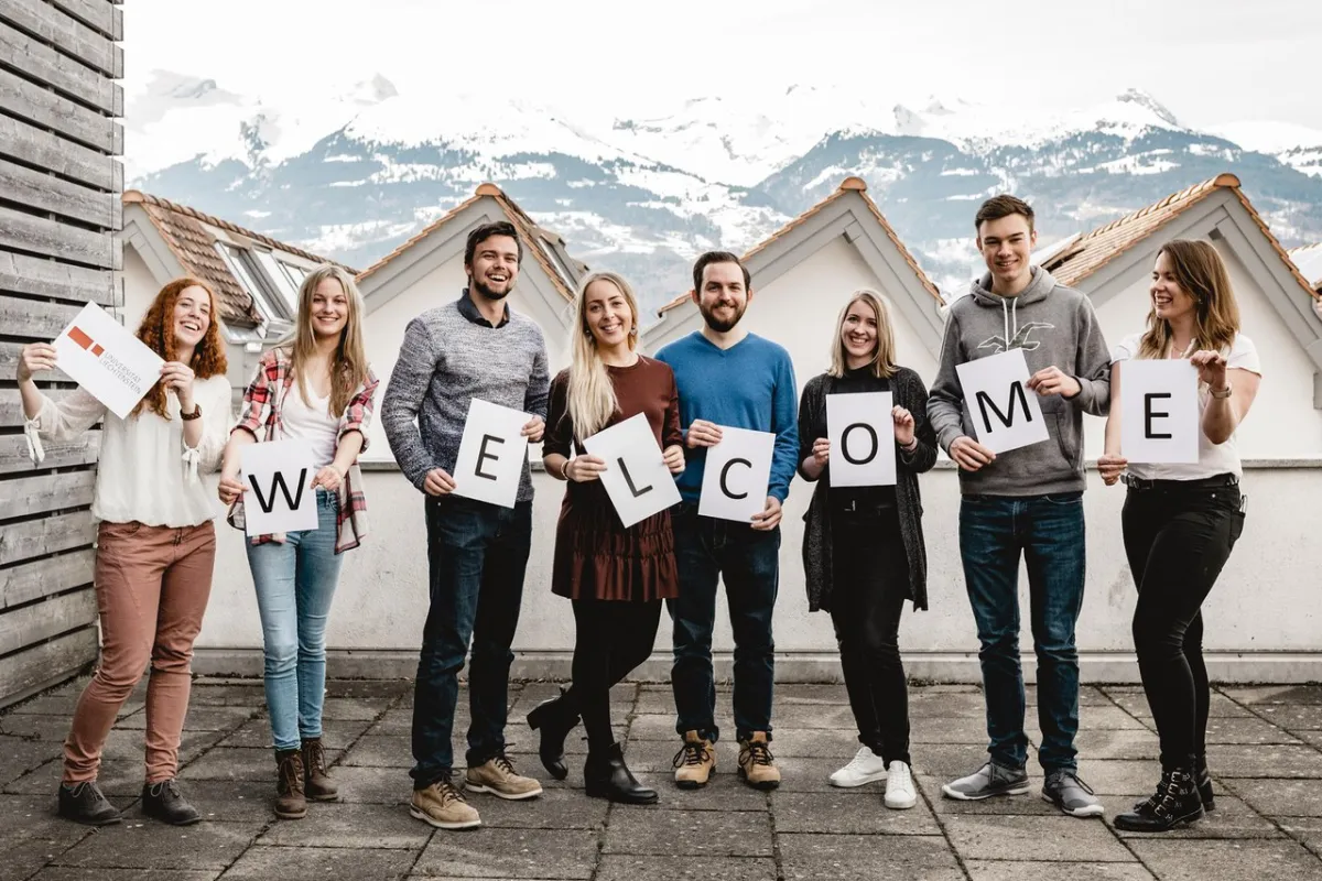 Eine Gruppe von acht jungen Menschen steht im Freien und hält große Buchstaben hoch, die das Wort "WELCOME" buchstabieren. Im Hintergrund sind Häuser mit Bergkulisse zu sehen, und die Gruppe lächelt in die Kamera, was eine freundliche und einladende Atmosphäre vermittelt.