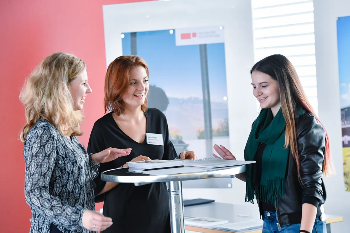 Drei Frauen im Gespräch an einem Stehtisch beim Infotag der Universität Liechtenstein, mit Informationsmaterialien und freundlicher Atmosphäre.