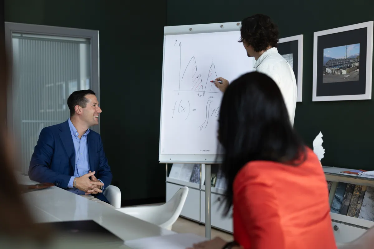 Teilnehmende bei einer Präsentation im Seminarraum, Fokus auf Vortragendem und Flipchart mit Funktionsgleichung.