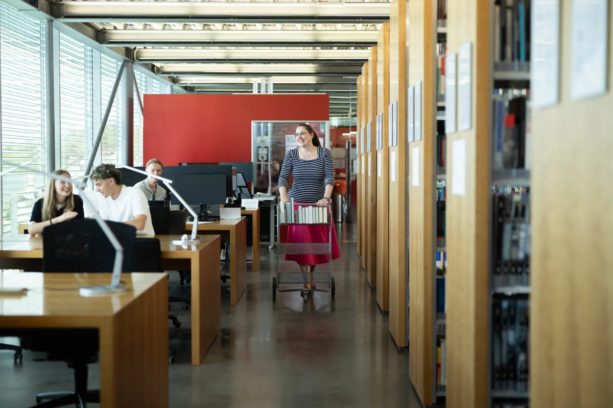 Studierende arbeiten und unterhalten sich in der Bibliothek, während die Bibliotheksmitarbeiterin mit Bücherwagen vorbeigeht.
