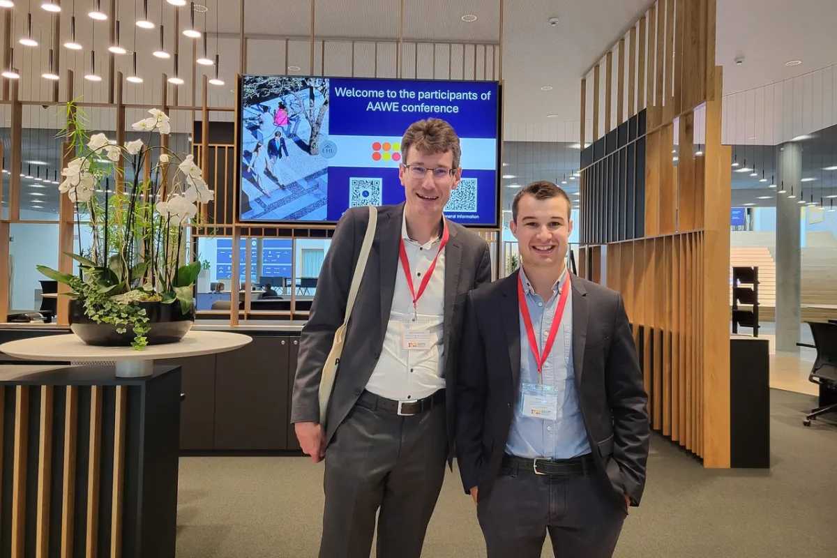Zwei Männer in Anzügen mit roten Namensschildern stehen lächelnd in einem modernen Konferenzraum. Im Hintergrund ist ein Bildschirm mit der Aufschrift „Welcome to the participants of AAWE conference“ zu sehen. Links steht eine dekorative Pflanze auf einem Tisch.