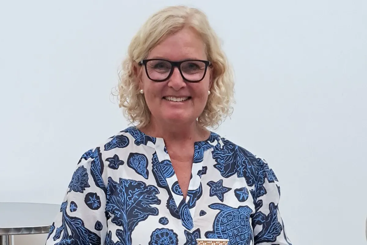 Eine lächelnde Frau mit blonden Haaren und Brille hält einen Becher in der Hand. Sie trägt ein blau-weiß gemustertes Kleid. Vor ihr steht ein Blumenstrauß in Folie verpackt.