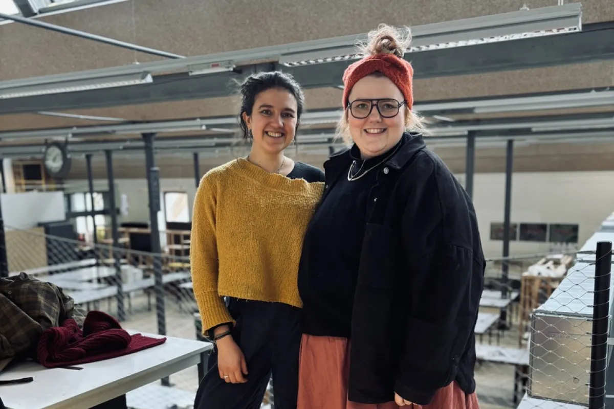 Zwei Personen stehen lächelnd in einem modernen Architekturatelier mit großen Fenstern und industriellem Design. Sie tragen lässige, kreative Kleidung. Im Hintergrund sind Arbeitsplätze und eine Uhr sichtbar.