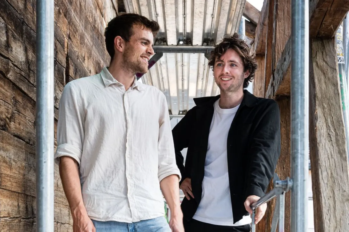 Zwei junge Männer stehen lächelnd auf einem Gerüst in einem Holzbau. Sie tragen legere Kleidung und wirken entspannt. Die Umgebung deutet auf eine Baustelle oder ein architektonisches Projekt hin.