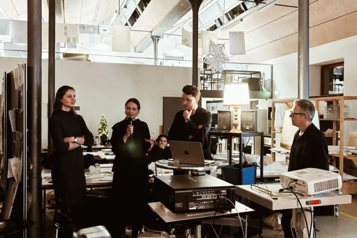 Eine Gruppe von Personen steht in einem modernen Architekturstudio. Eine Person spricht in ein Mikrofon, während andere aufmerksam zuhören. Im Raum sind technische Geräte und Präsentationsmaterialien zu sehen.