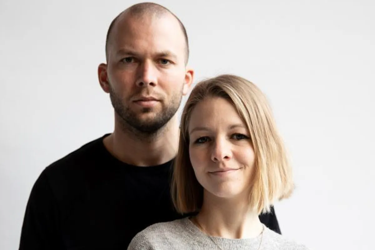 Ein Mann und eine Frau stehen vor einem neutralen Hintergrund. Beide blicken direkt in die Kamera. Die Frau trägt einen grauen Pullover, der Mann ein schwarzes T-Shirt.