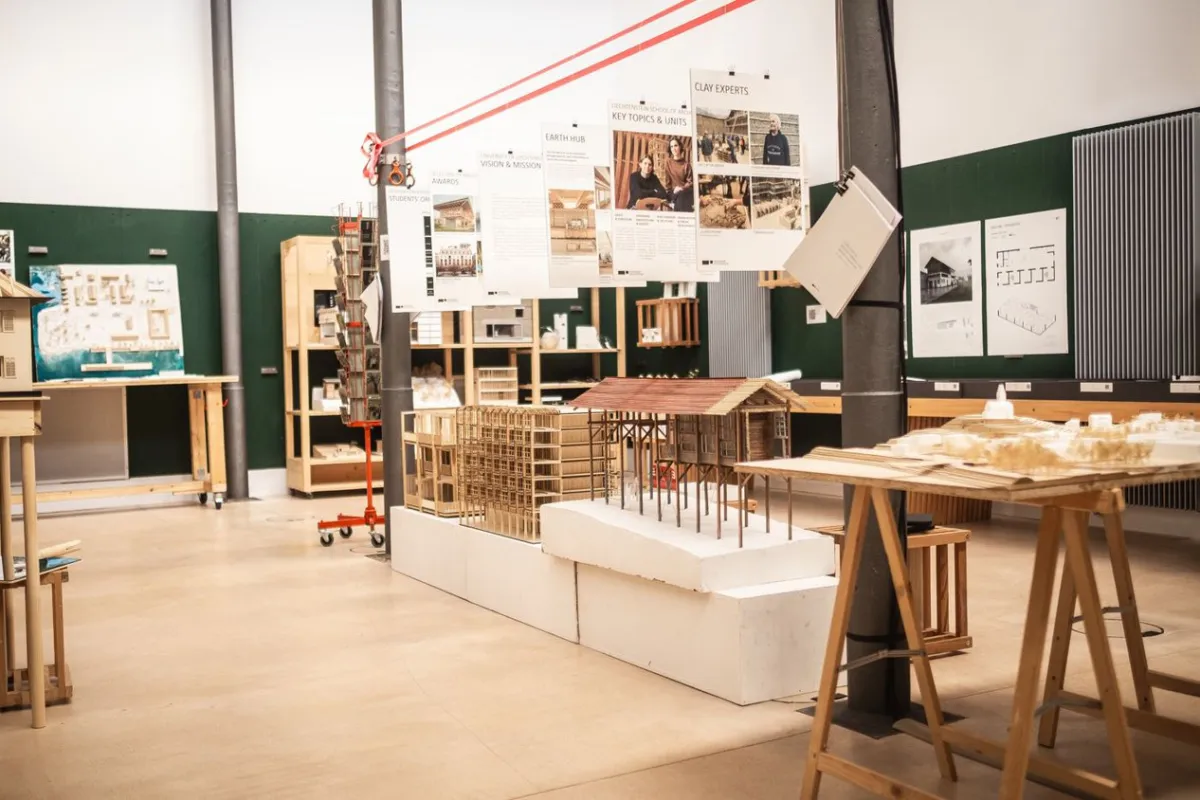 Architektur-Studienraum mit Modellen und Plakaten. Verschiedene Architekturmodelle aus Holz und Papier sind auf Tischen ausgestellt. An den Wänden hängen Plakate mit Informationen zu Projekten und Themen. Der Raum ist hell und modern gestaltet.