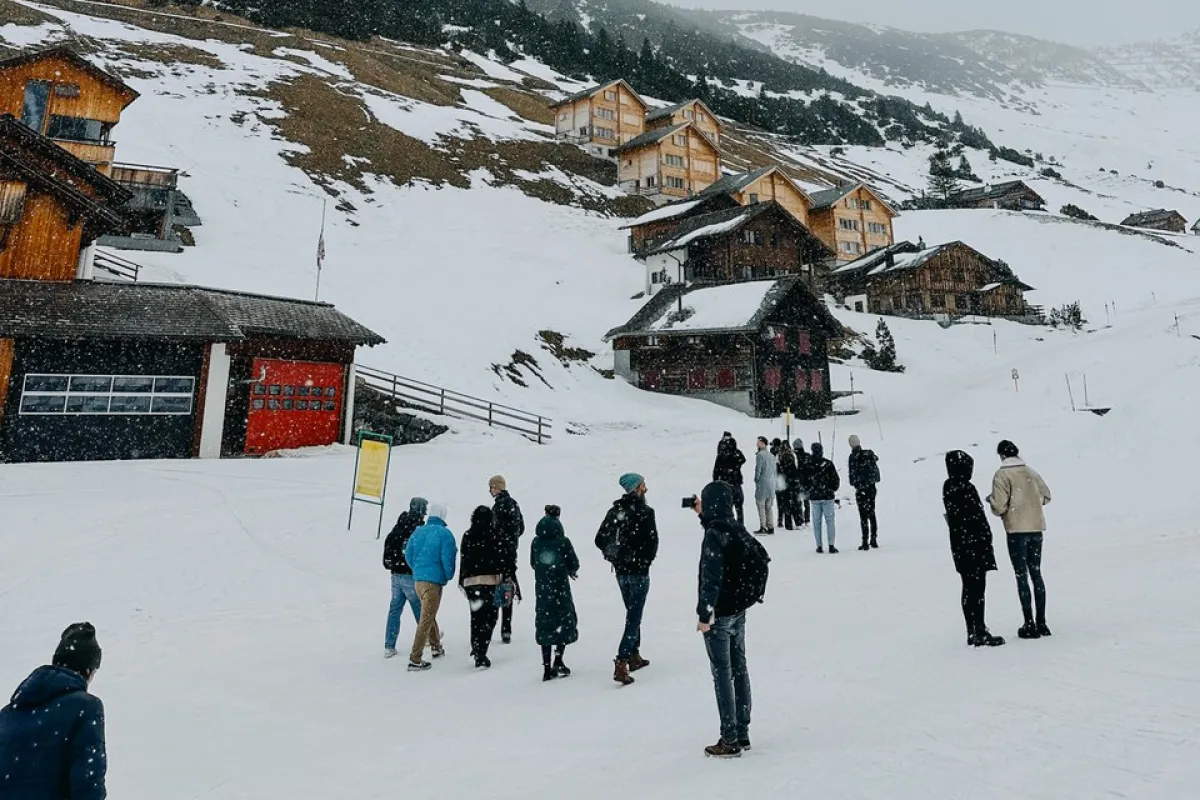 Eine Gruppe von Menschen steht in einer verschneiten Berglandschaft vor traditionellen Holzhäusern. Im Hintergrund sind schneebedeckte Hügel und Nadelbäume zu sehen. Die Szene vermittelt eine winterliche Atmosphäre und zeigt eine Verbindung von Architektur und Natur.