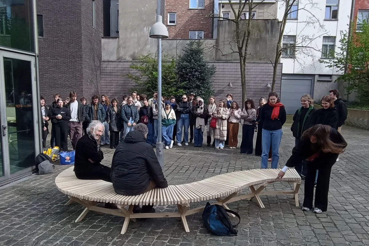 Eine Gruppe von Studierenden steht im Freien um eine geschwungene Holzbank, die von zwei Personen genutzt wird. Die Szene findet auf einem gepflasterten Platz vor modernen Gebäuden statt.