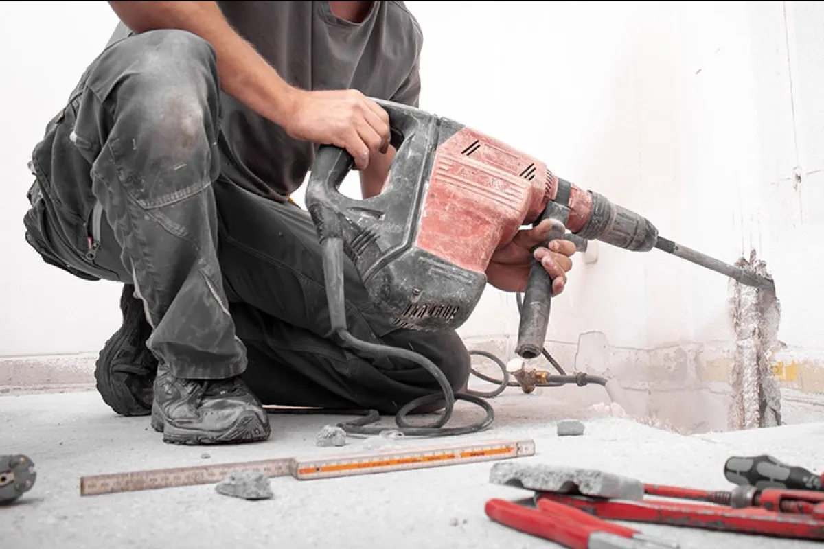 Ein Bauarbeiter kniet auf dem Boden und benutzt einen roten Kombi-Hammer, um eine Wand zu bearbeiten. Im Vordergrund liegen Werkzeuge und Bauutensilien.