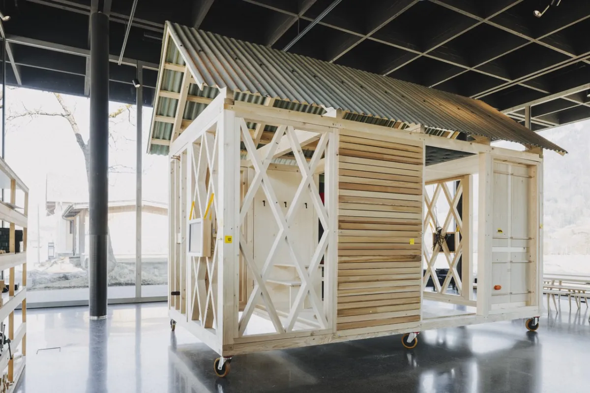 Ein einfaches, modulares Holzgebäude mit einem Wellblechdach steht auf Rollen in einem modernen, lichtdurchfluteten Raum. Das Design zeigt eine offene Struktur mit diagonalen Verstrebungen und Holzverkleidung, ideal für nachhaltiges Bauen und architektonische Bildung.