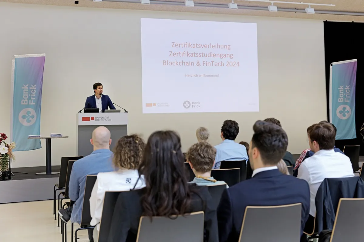 Ein Redner steht an einem Podium vor einer Leinwand mit der Aufschrift "Zertifikatsverleihung Zertifikatsstudiengang Blockchain & FinTech 2024" bei der Universität Liechtenstein. Im Publikum sitzen mehrere Personen. Links und rechts sind Banner von Bank Frick zu sehen.