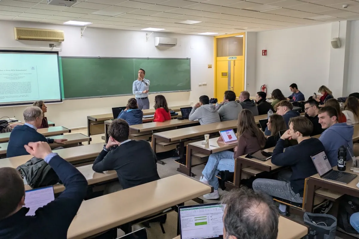 Ein Dozent hält einen Vortrag in einem Hörsaal, umgeben von Studierenden, die aufmerksam zuhören und Notizen auf Laptops machen. Im Hintergrund ist eine Tafel und eine Präsentation zu sehen.