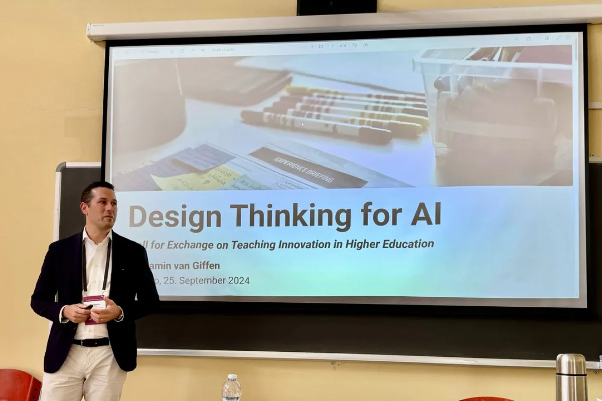 Ein Mann steht vor einer Leinwand, auf der eine Präsentation mit dem Titel "Design Thinking for AI" gezeigt wird. Der Raum wirkt wie ein Seminarraum, und es sind einige Stifte und Notizen auf der Leinwand zu sehen.