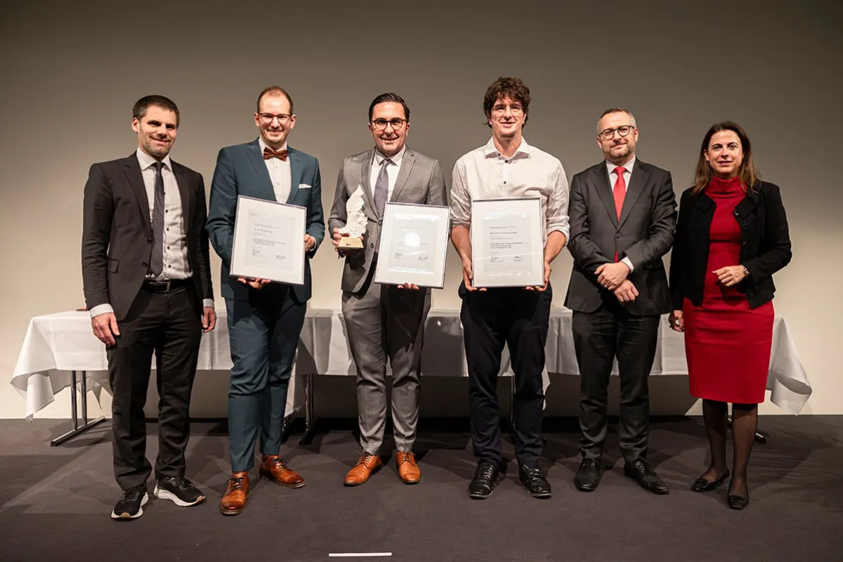 Eine Gruppe von sechs Personen steht nebeneinander auf einer Bühne. Drei Personen in der Mitte halten Auszeichnungen und Urkunden. Alle sind formell gekleidet und lächeln in die Kamera. Im Hintergrund sind weiße Tischdecken sichtbar.