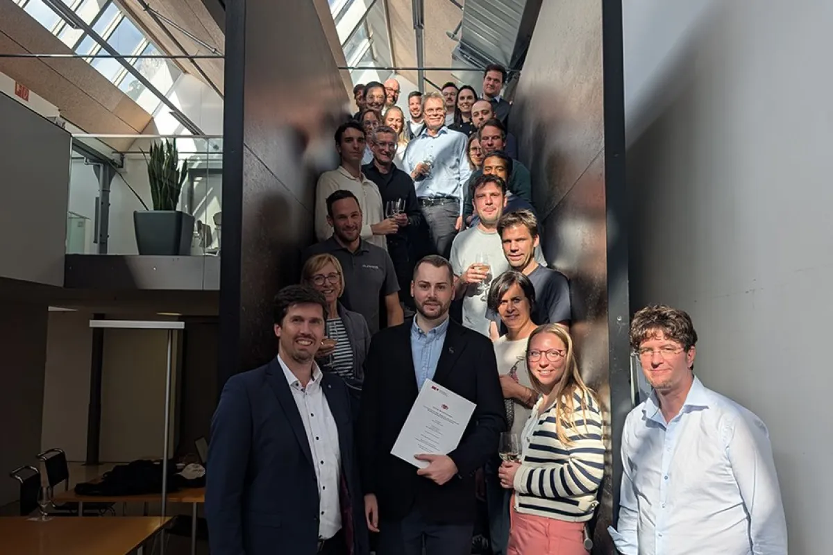 Eine Gruppe von Menschen steht auf einer Treppe in einem modernen Gebäude. Im Vordergrund hält eine Person ein Dokument in der Hand, umgeben von lächelnden Kollegen. Die Szene ist gut beleuchtet durch ein Oberlicht.