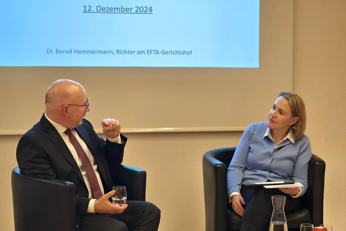 Zwei Personen sitzen in Sesseln vor einer Leinwand, die das Datum "12. Dezember 2024" zeigt. Eine Person hält ein Glas Wasser, die andere hat ein Notizbuch auf dem Schoß. Im Vordergrund steht eine Karaffe auf einem Tisch mit weihnachtlicher Dekoration.