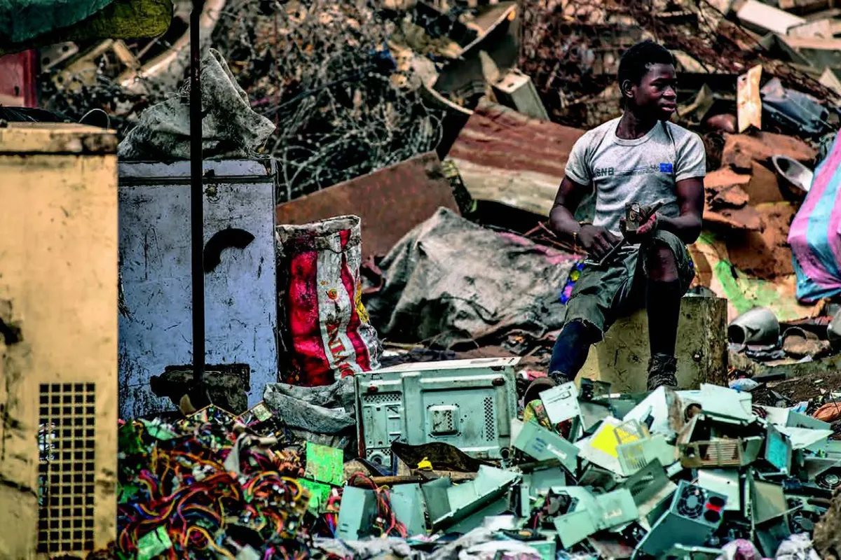 Ein Mann sitzt auf einem Hocker inmitten von Elektroschrott auf einer Müllhalde. Um ihn herum sind alte Geräte und Kabel zu sehen. Die Szene zeigt die Problematik von Elektroschrott und dessen Auswirkungen auf die Umwelt.