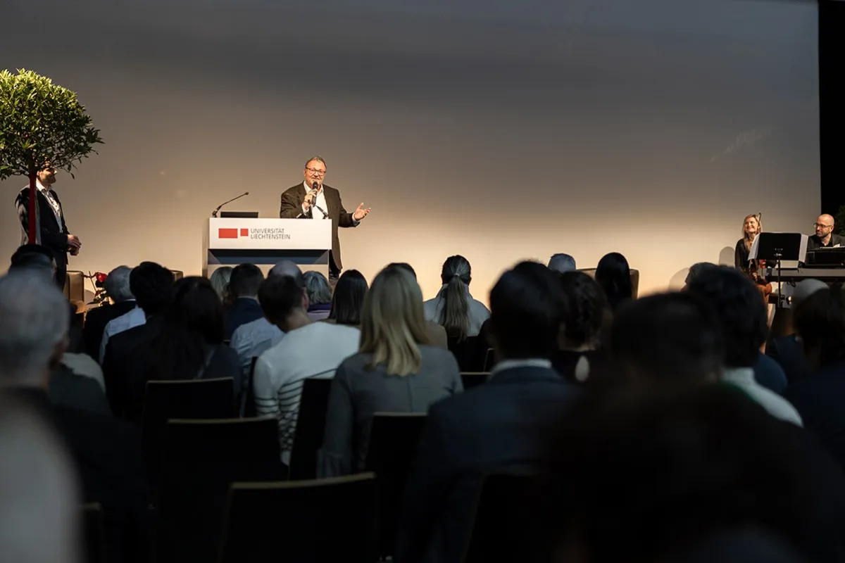Ein Redner steht an einem Podium mit dem Logo der Universität Liechtenstein, spricht zu einem Publikum. Im Hintergrund sind zwei Musiker zu sehen. Die Veranstaltung findet in einem großen Saal statt.
