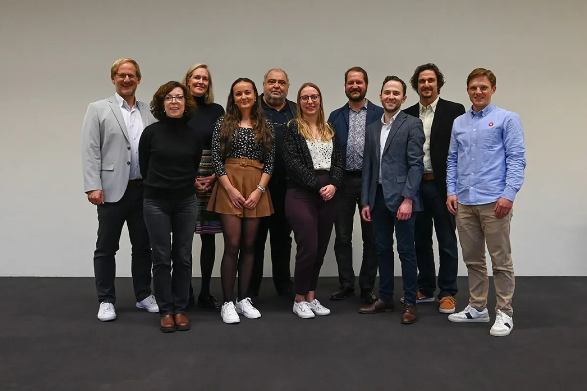 Eine Gruppe von zehn Personen steht lächelnd nebeneinander vor einem neutralen Hintergrund. Sie tragen unterschiedliche, teils formelle, teils legere Kleidung. Das Bild vermittelt eine freundliche und professionelle Atmosphäre.