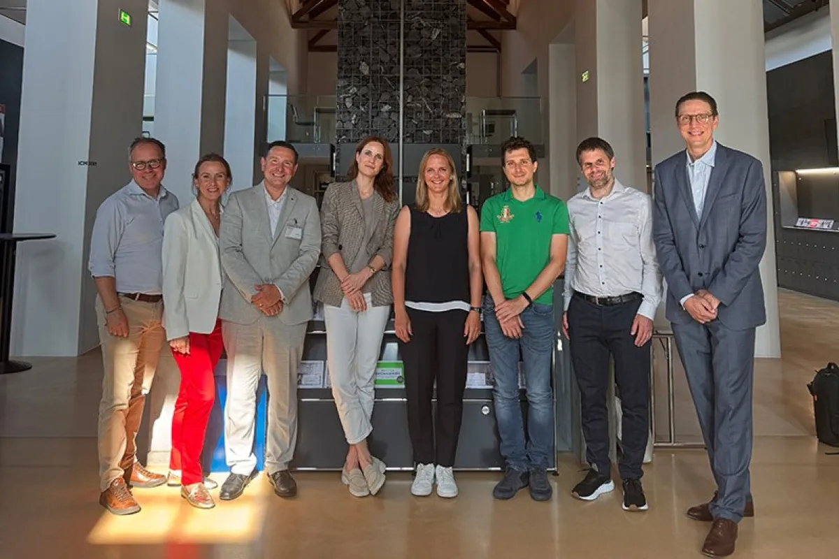Eine Gruppe von neun Personen steht in einer modernen Halle mit Holzdach und einem Kronleuchter. Sie lächeln in die Kamera. Im Hintergrund sind hohe Fenster und eine Steinwand zu sehen.