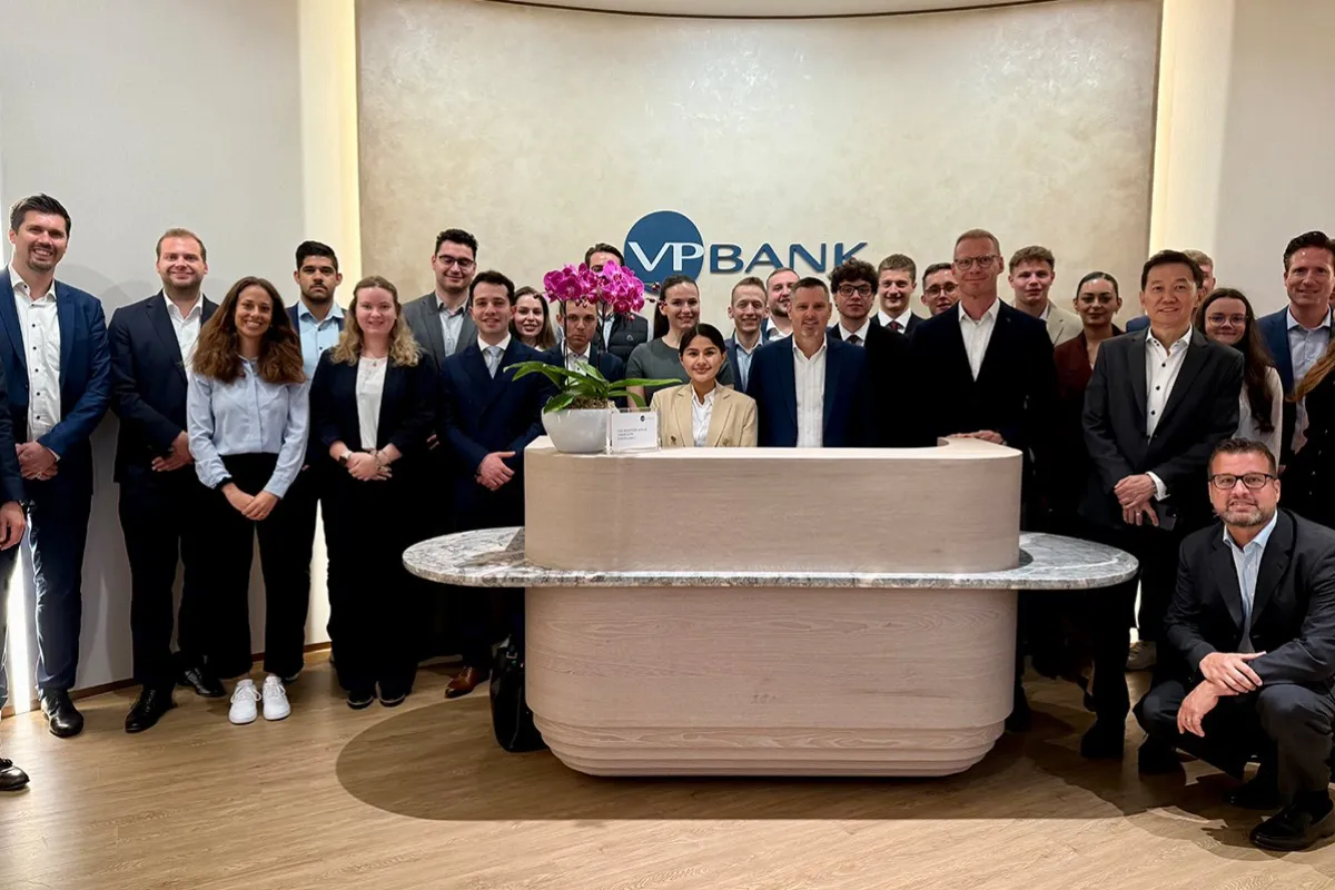 Gruppe von Studierenden und Fachleuten in formeller Kleidung steht in der Lobby der VP Bank. Im Hintergrund ist das Logo der Bank sichtbar. Die Szene vermittelt eine professionelle und lernorientierte Atmosphäre.