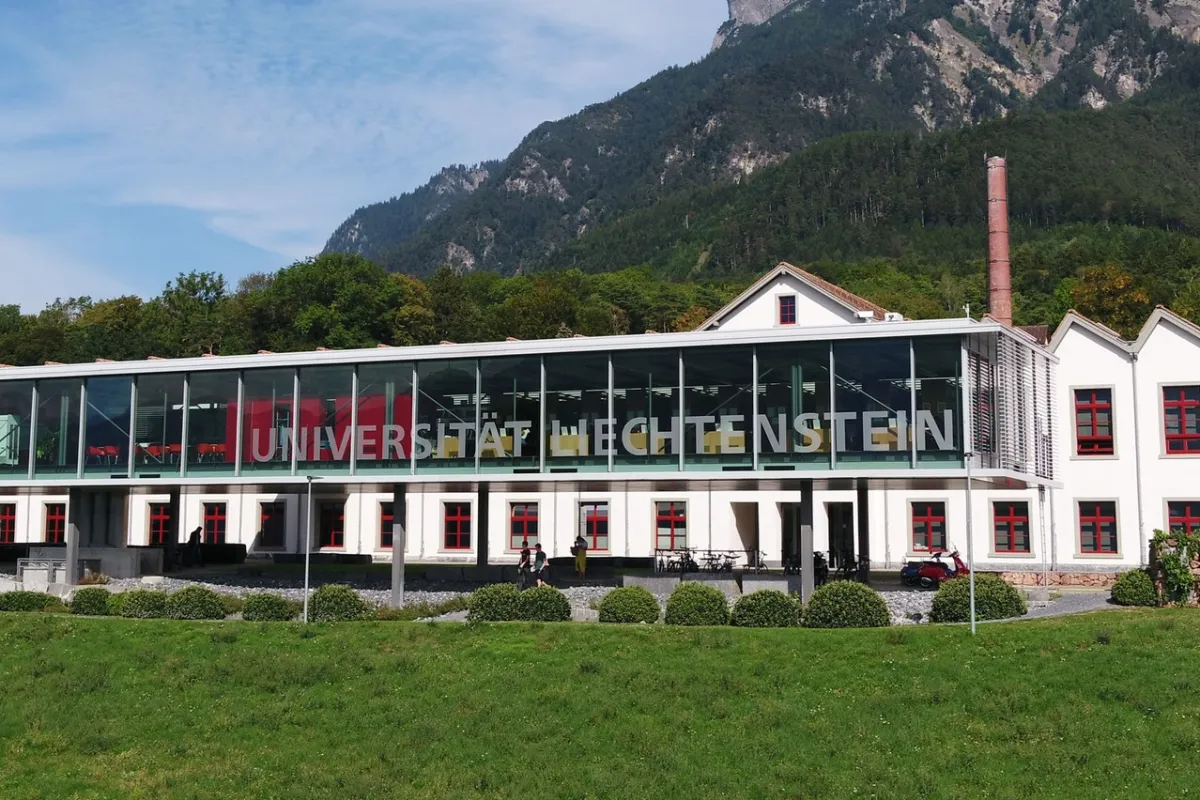 Außenansicht der Universität Liechtenstein mit modernem Glasgebäude und traditionellem weißen Gebäude im Hintergrund, umgeben von grüner Landschaft und Bergen.