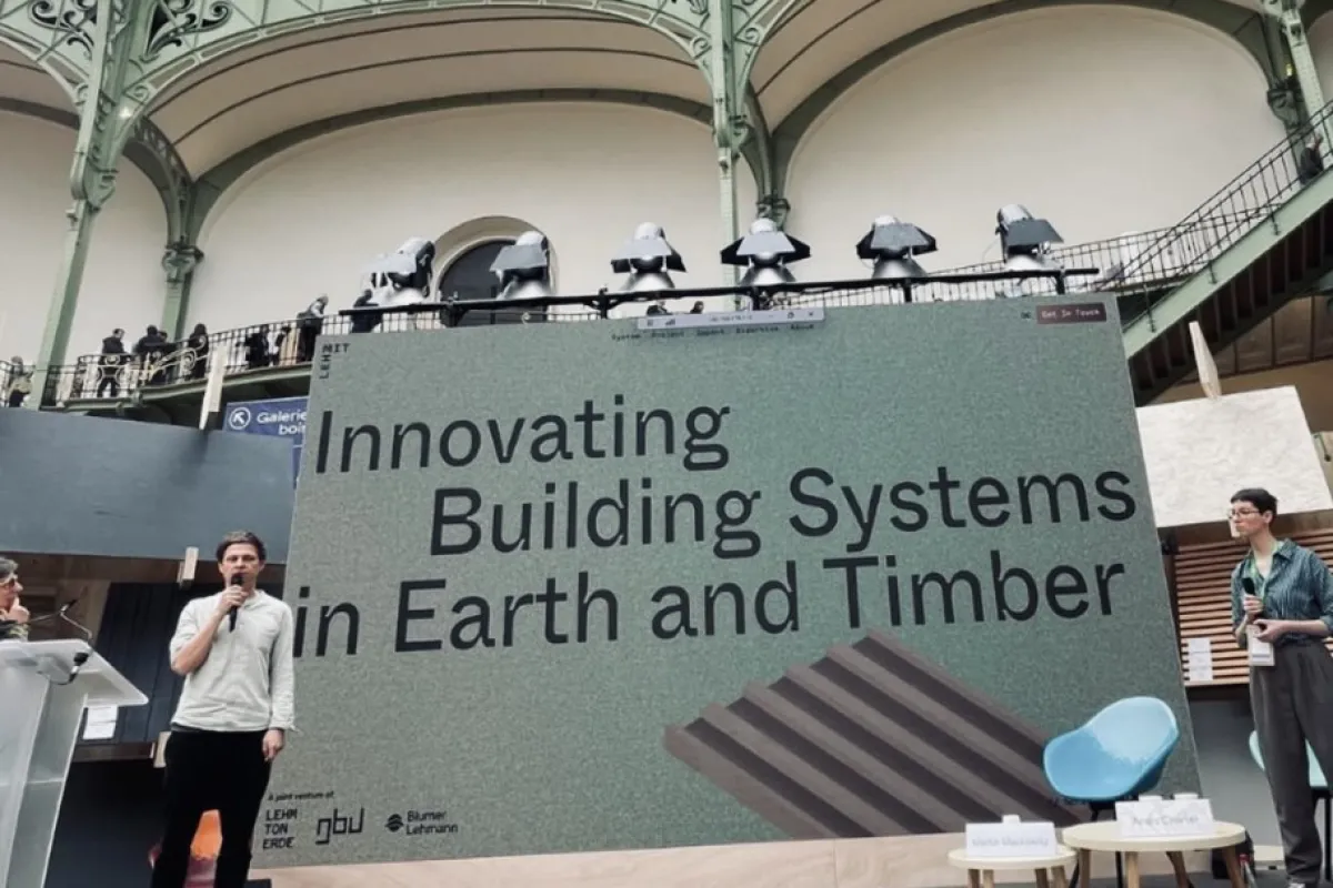 Ein Redner präsentiert auf einer Bühne vor einem großen Bildschirm mit der Aufschrift „Innovating Building Systems in Earth and Timber“ im Grand Palais, Paris. Im Hintergrund ist die beeindruckende Architektur des Veranstaltungsortes zu sehen.