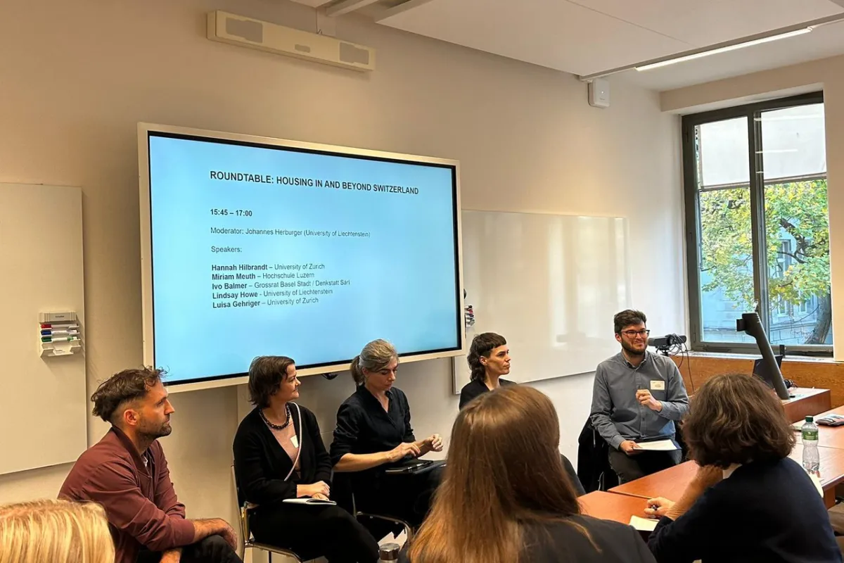Eine Gruppe von Personen sitzt in einem Seminarraum und diskutiert bei einer Podiumsdiskussion zum Thema "Housing in and Beyond Switzerland". Im Hintergrund ist ein Bildschirm mit den Namen der Teilnehmenden und dem Moderator zu sehen.