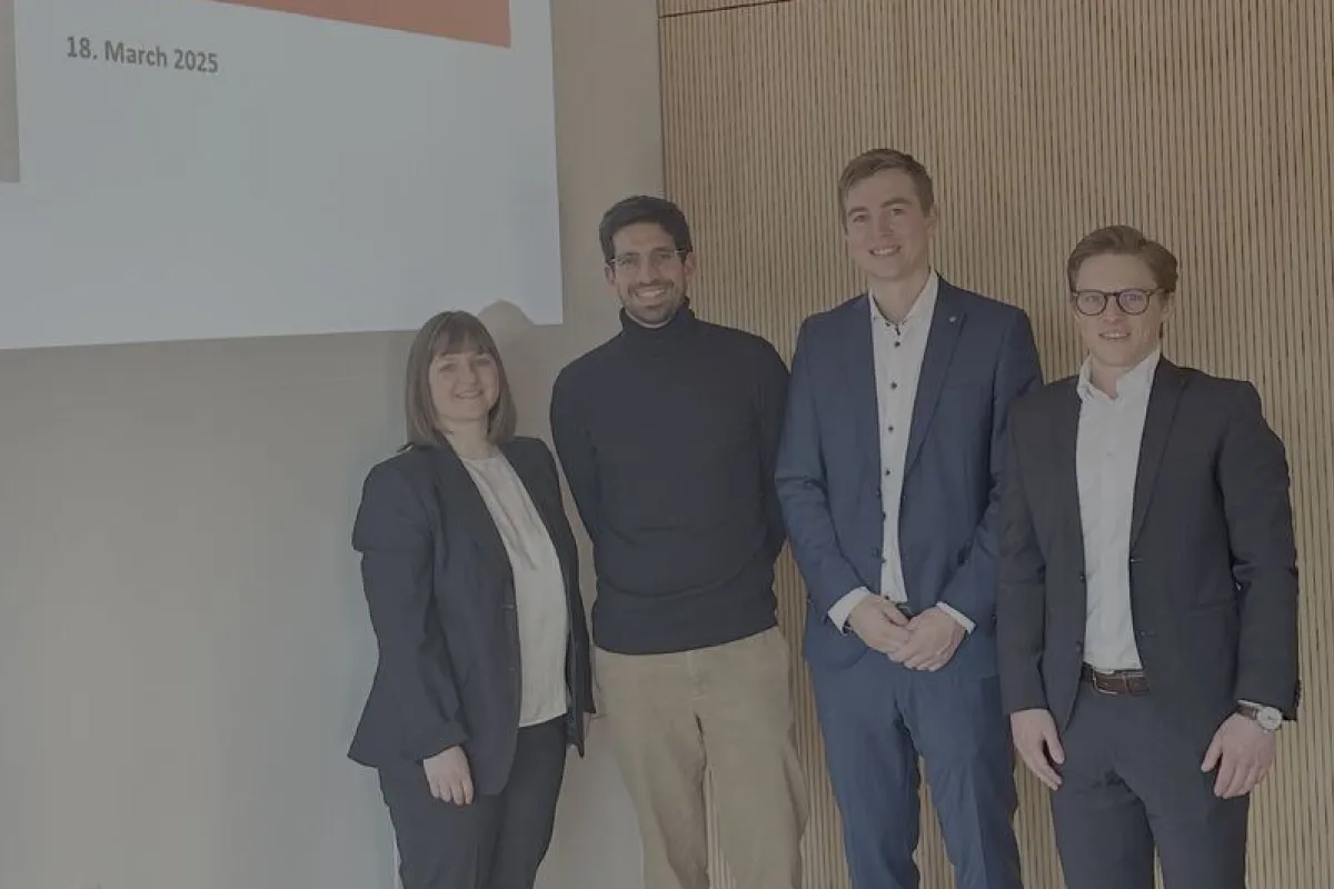 Vier Personen stehen lächelnd vor einer Präsentationsfolie mit der Aufschrift "LAB: Sustainability, Prosperity, and Provision, 18. March 2025". Sie befinden sich in einem modernen Raum mit Holzvertäfelung.