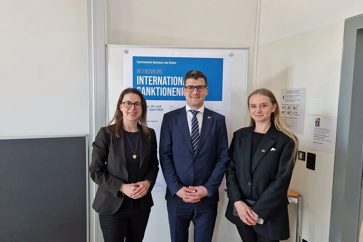Drei Personen stehen vor einem Poster mit der Aufschrift "Intensivkurs Internationales Sanktionenrecht". Sie tragen formelle Kleidung und lächeln in die Kamera.