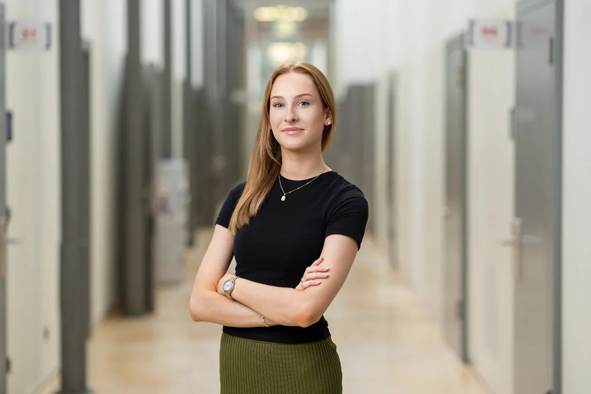 Eine junge Frau steht selbstbewusst mit verschränkten Armen in einem modernen Flur. Sie trägt ein schwarzes Oberteil und einen grünen Rock.