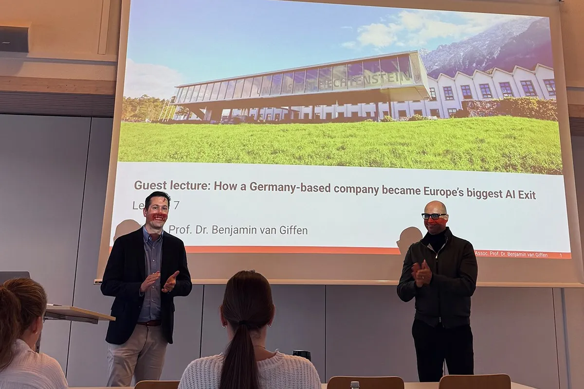 Zwei Männer stehen vor einer Präsentationsfolie in einem Vorlesungssaal der Universität Liechtenstein. Die Folie zeigt den Titel eines Gastvortrags über den Erfolg eines deutschen KI-Unternehmens. Studierende sitzen im Publikum.