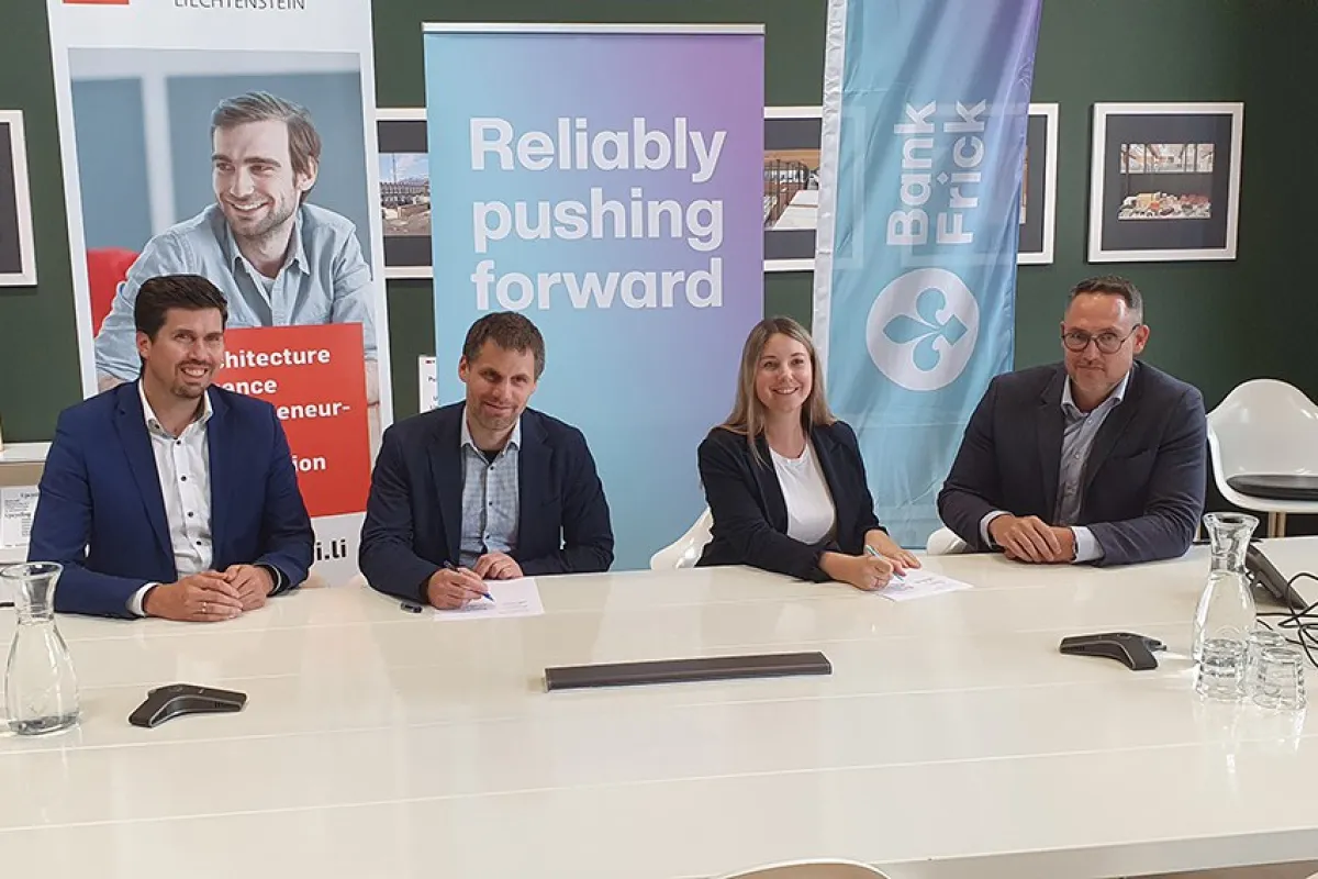 Vier Personen sitzen an einem Konferenztisch und unterzeichnen Dokumente. Im Hintergrund sind Banner der Universität Liechtenstein und Bank Frick zu sehen. Die Szene symbolisiert die Verlängerung der Kooperation zwischen der Universität und der Bank.