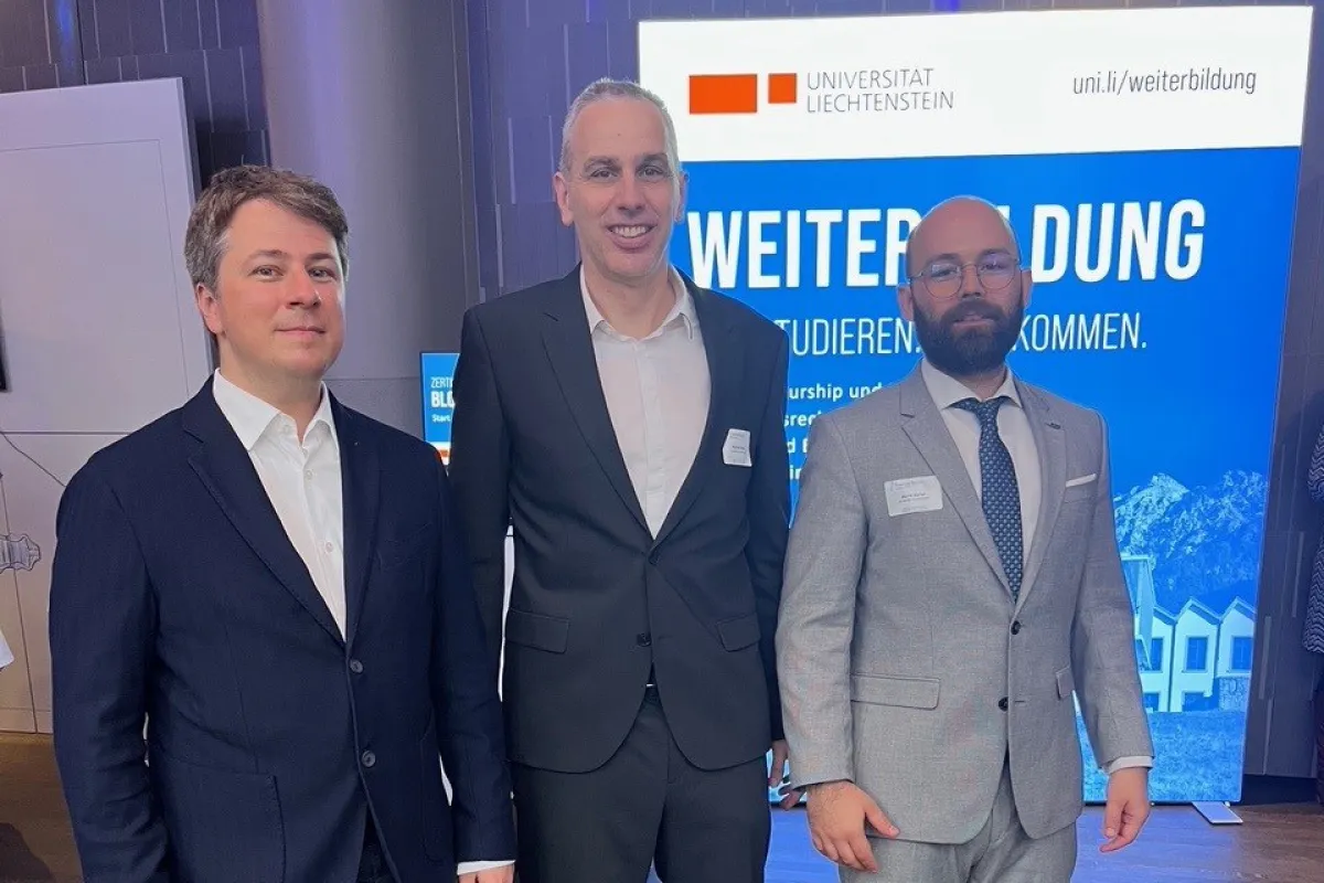 Drei Männer in Anzügen stehen vor einem Schild der Universität Liechtenstein. Im Hintergrund ist ein Banner mit der Aufschrift "Weiterbildung" zu sehen.