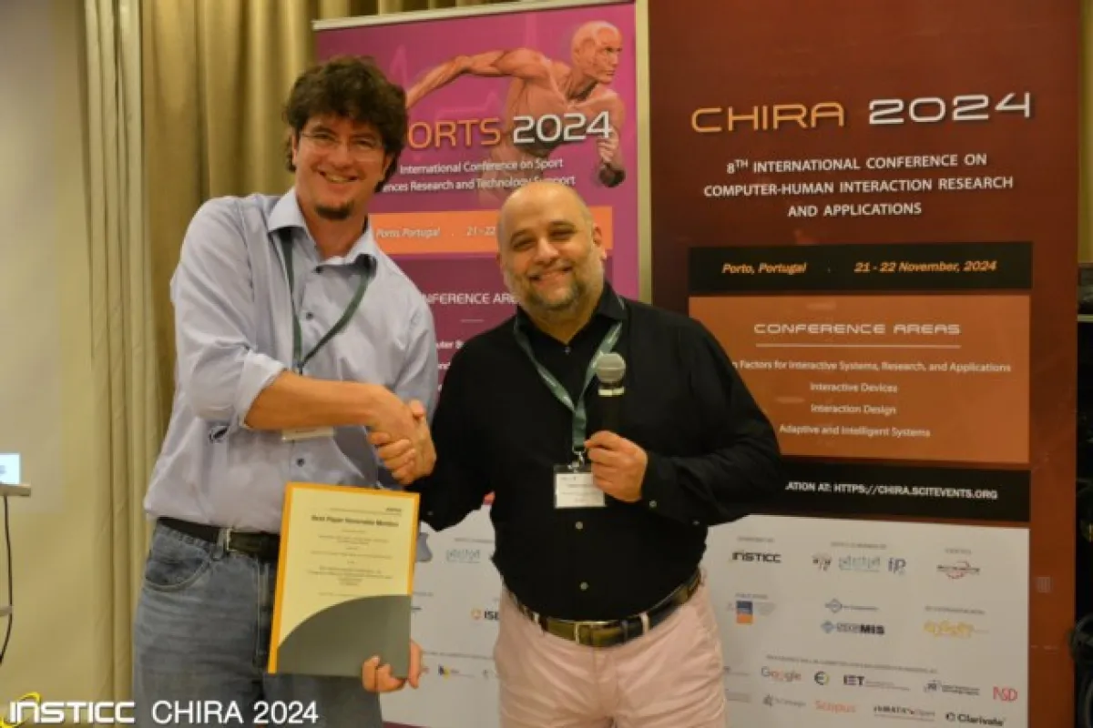 Zwei Männer stehen vor Bannern der CHIRA 2024 Konferenz. Einer hält ein Zertifikat, während der andere ein Mikrofon hält. Beide lächeln und schütteln sich die Hand. Im Hintergrund sind Details zur Konferenz in Porto, Portugal, sichtbar.