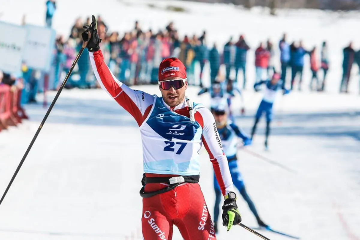 Langläufer überquert bei sonnigem Winterwetter als Sieger die Ziellinie eines Langlaufrennens, trägt einen rot-weißen Rennanzug mit Startnummer 27, hebt den Arm zum Jubel – im Hintergrund jubelnde Zuschauer und schneebedeckte Landschaft.