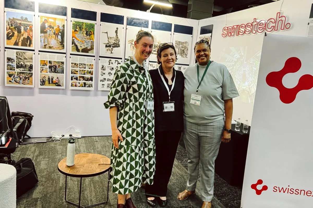 Drei Personen stehen lächelnd vor Ausstellungstafeln mit Bildern und Texten. Im Hintergrund ist ein Banner mit dem Logo von "swisstech" zu sehen. Die Szene befindet sich in einem Ausstellungsraum, der Teil der Präsentation "Making Urban Africa" auf dem South African Science Forum ist.