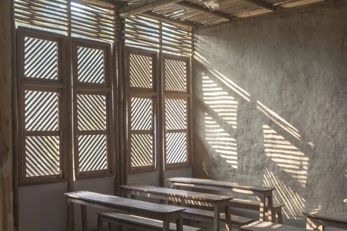 Innenraum eines nachhaltig gebauten Klassenzimmers mit Holzbänken und Wänden aus natürlichen Materialien. Sonnenlicht fällt durch hölzerne Fensterläden und erzeugt Muster auf der Wand.