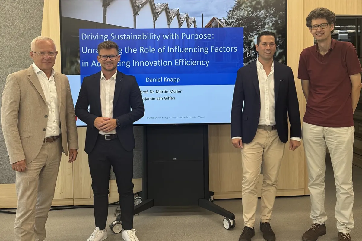 Vier Männer stehen vor einem Bildschirm, der eine Präsentation mit dem Titel „Driving Sustainability with Purpose“ zeigt. Sie tragen Business-Kleidung und lächeln in die Kamera. Der Hintergrund ist ein moderner Innenraum.