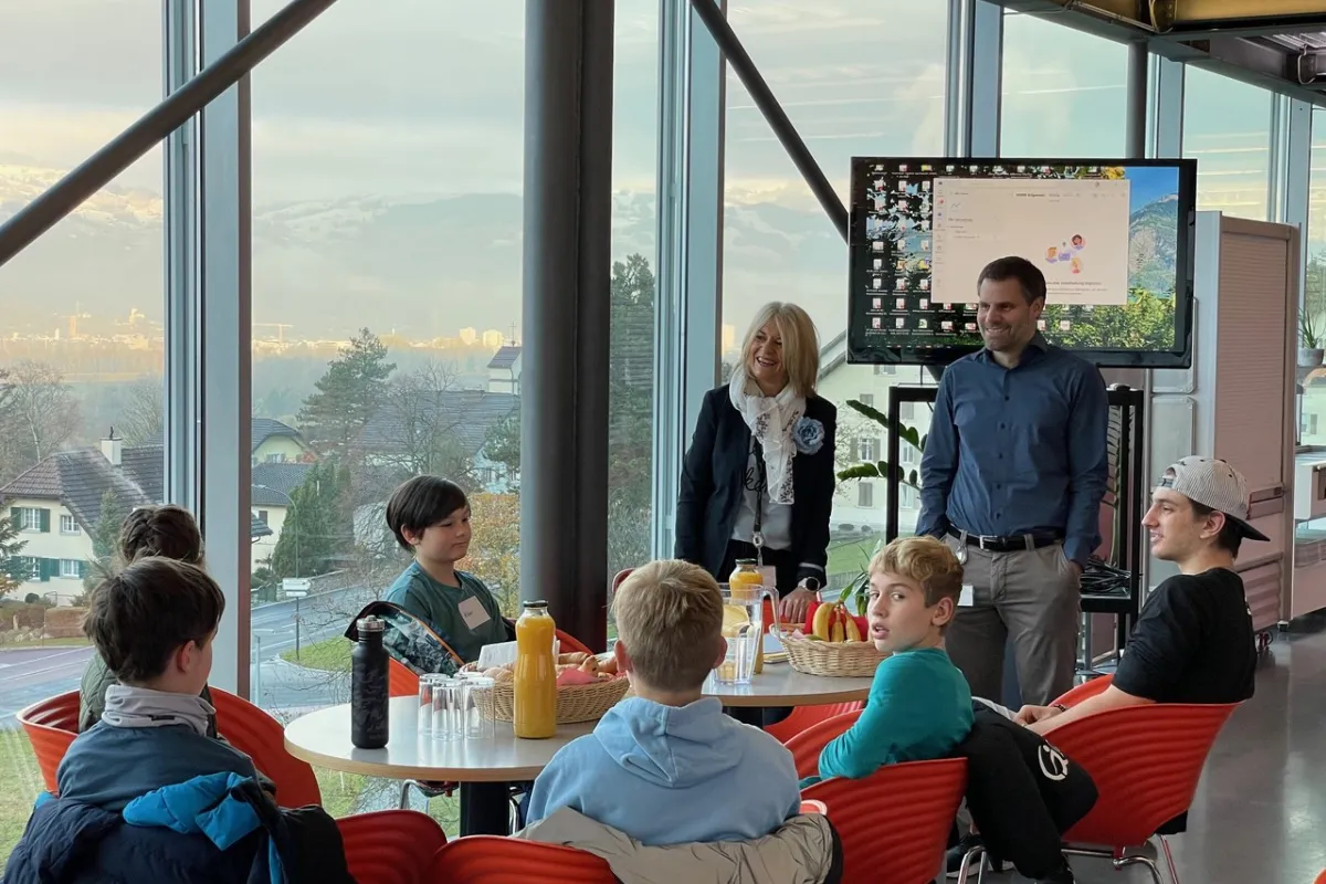 Eine Gruppe von Kindern sitzt an einem Tisch in einem modernen Raum mit großen Fenstern. Zwei Erwachsene stehen lächelnd daneben. Im Hintergrund ist ein Bildschirm zu sehen. Die Szene wirkt freundlich und einladend.