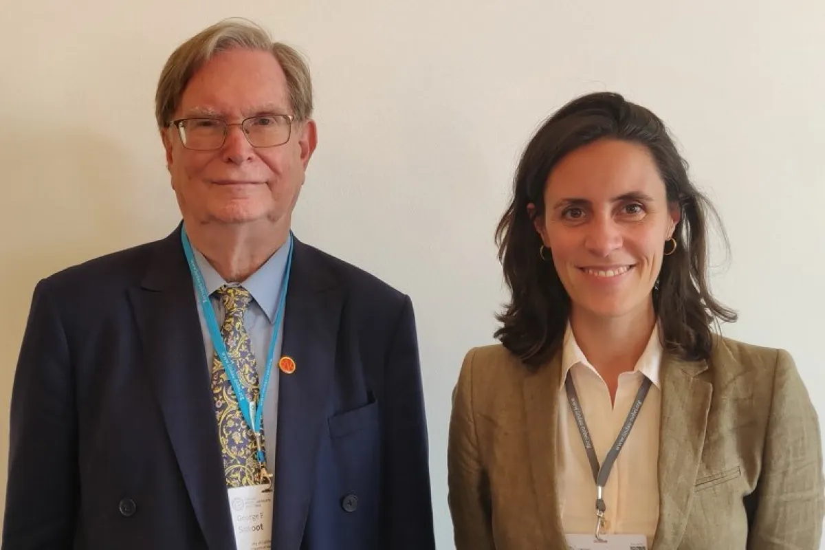 Zwei Personen stehen nebeneinander und lächeln in die Kamera. Beide tragen Namensschilder und sind formell gekleidet.