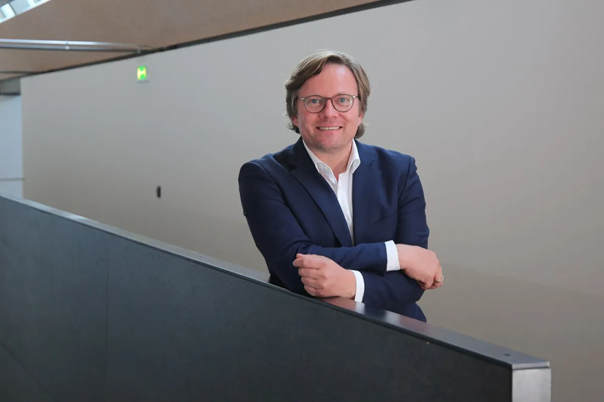 Ein Mann in einem blauen Anzug lehnt lächelnd an einem Geländer in einem modernen Gebäude.